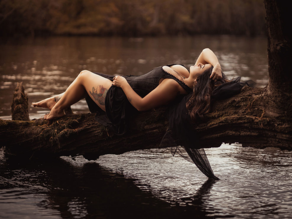 A portrait of a beautiful woman in a black corset laying on a tree branch by the Black River in NC captured by Salty Star Photography