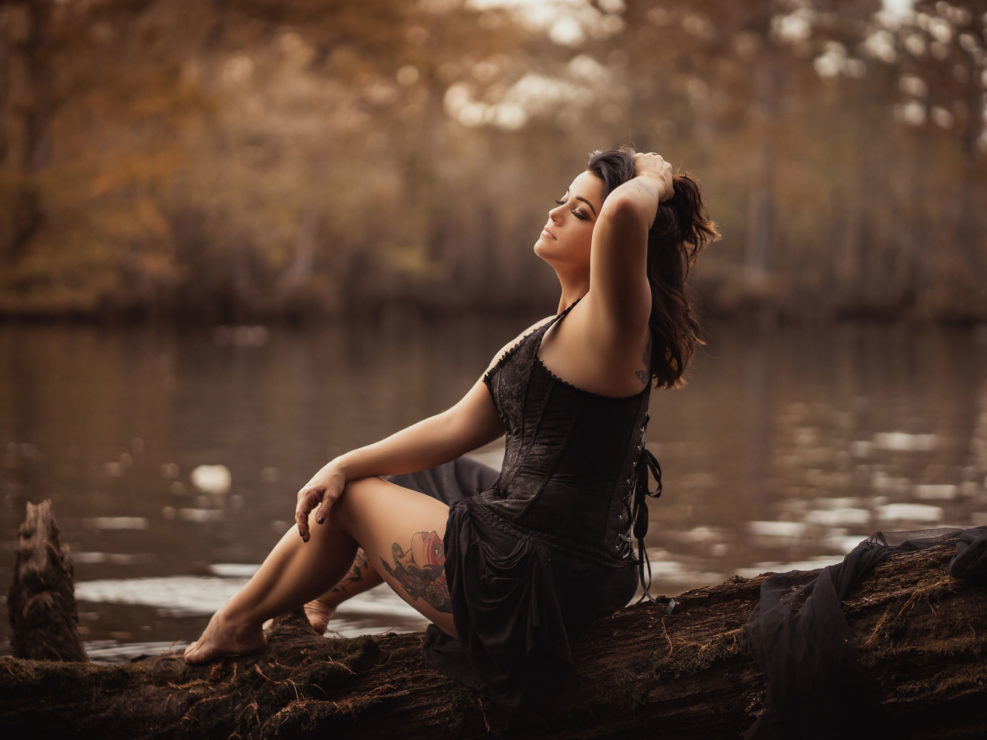 A portrait of a beautiful woman in a black corset sitting on a tree branch by the Black River in NC captured by Salty Star Photography